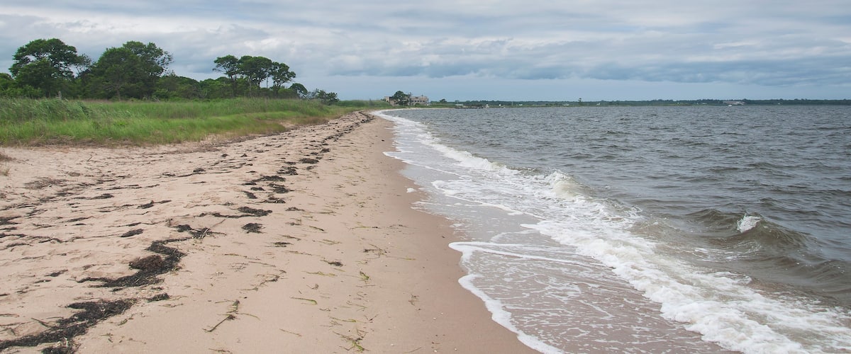 A sandy beach with gentle waves, grassy dunes, and a cloudy sky.