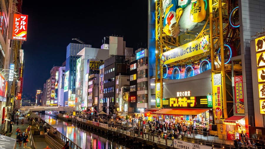 A bustling nighttime street with illuminated signs and a river in the foreground.
