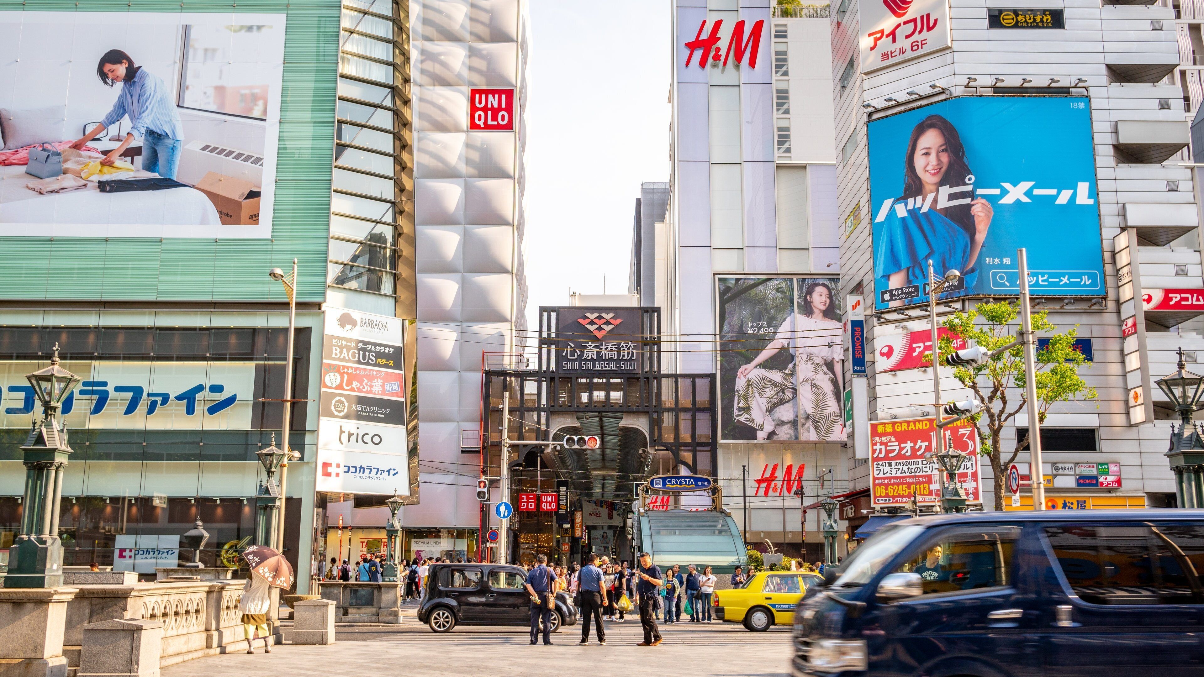Eine belebte Stadtstraße mit verschiedenen Geschäftsgebäuden, darunter ein großes H&M Geschäft und eine Werbetafel, die ein Schönheitsprodukt bewirbt.