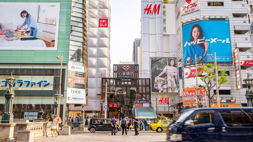 Eine belebte Stadtstraße mit verschiedenen Geschäftsgebäuden, darunter ein großes H&M Geschäft und eine Werbetafel, die ein Schönheitsprodukt bewirbt.