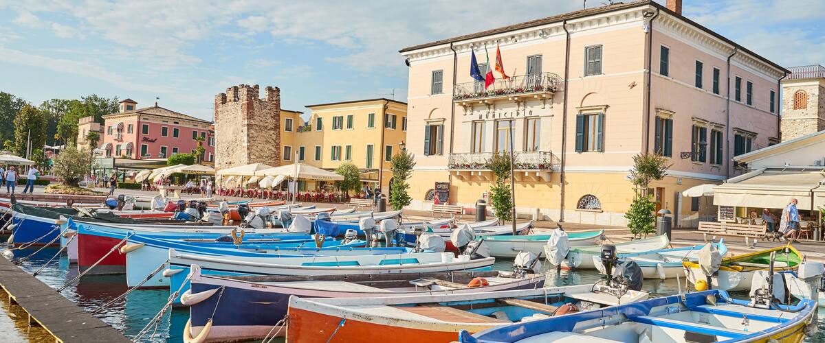 A marina with colorful boats docked, a historic building with flags, and a castle in the background.