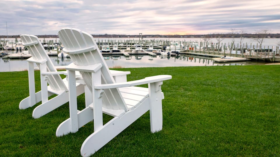 Three white Adirondack chairs on a grassy lawn with a marina and boats in the background.