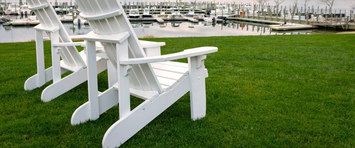 White "Adirondack" style lounge chairs on green grass at waterfront marina, with boats docked in background