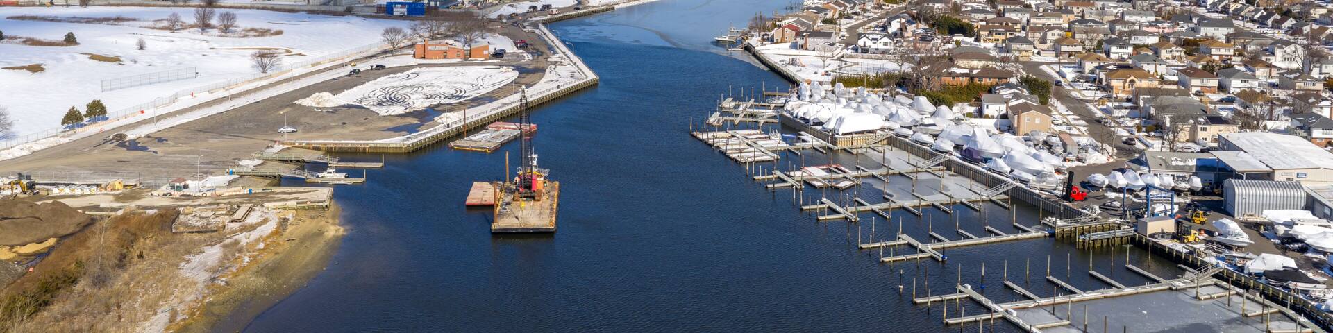 Aerial view of the oceanside waterway and a winter marina with empty boat slips, surrounded by snow covered residential neighborhoods and commercial areas in nassau county, new york