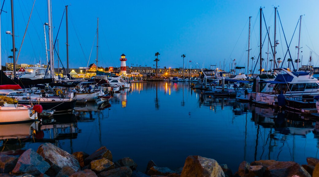 Un puerto deportivo al anochecer con barcos amarrados, un faro destacado y un litoral rocoso.
