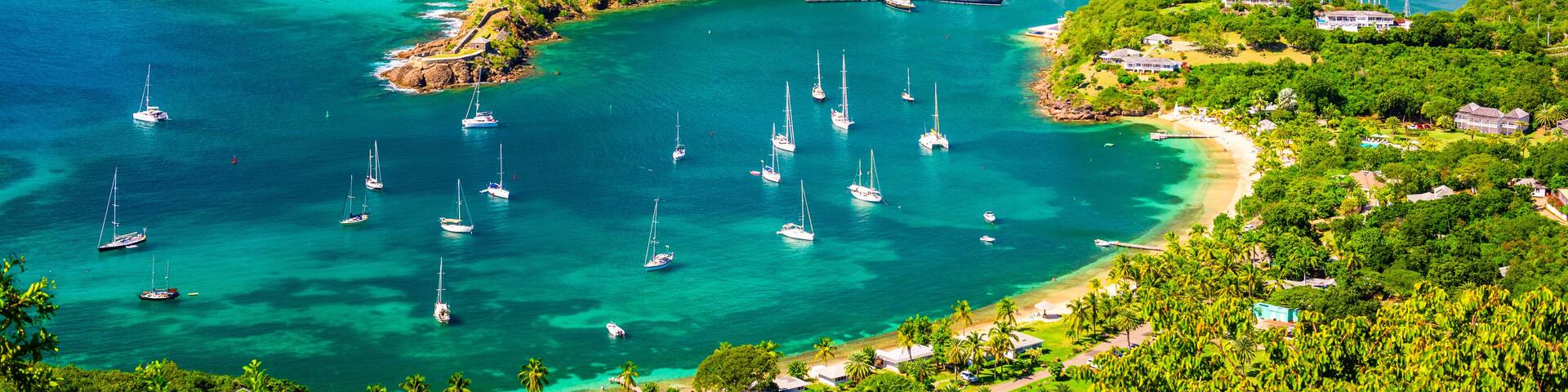 A coastal landscape with clear turquoise waters, numerous sailboats, lush green hills, and a small harbor with buildings.