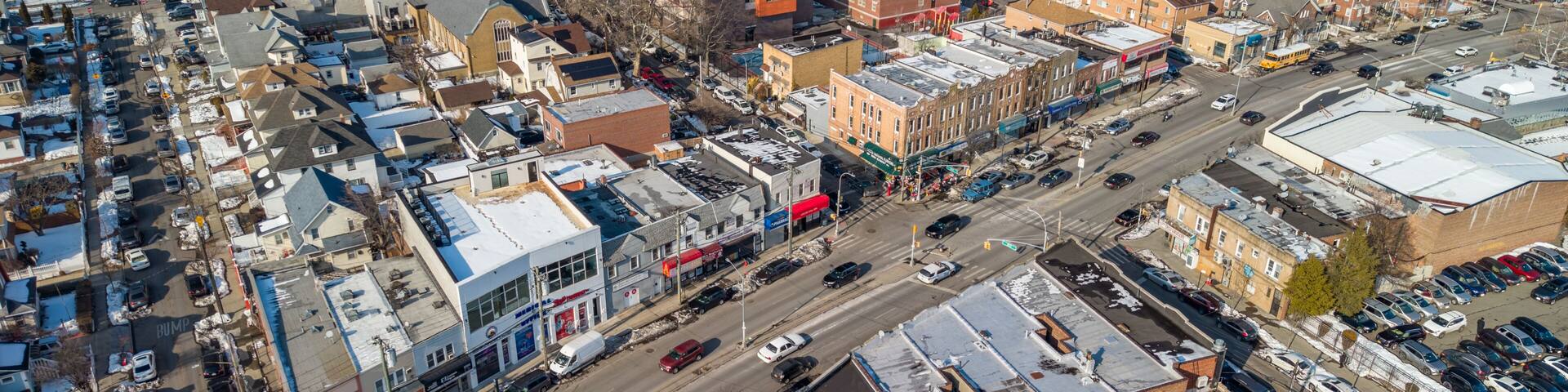 Hollis queens neighborhood overview portraying a bustling street with cars, diverse residential and commercial buildings under a clear sky during winter with snow patches