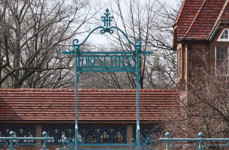 Una reja de hierro forjado con la inscripción 'FOREST HILLS', un edificio de ladrillo y árboles sin hojas al fondo.