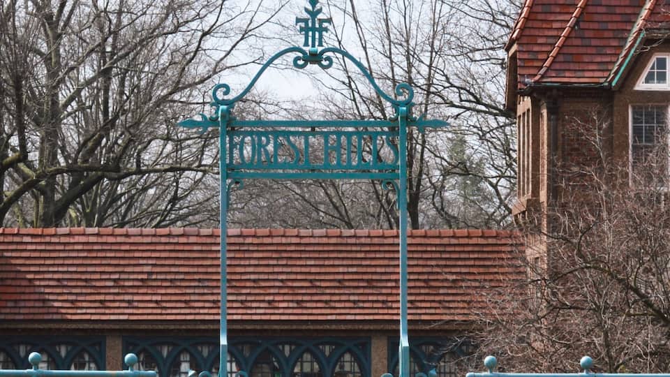 A wrought iron gate with 'FOREST HILLS' inscribed, a brick building, and leafless trees in the background.