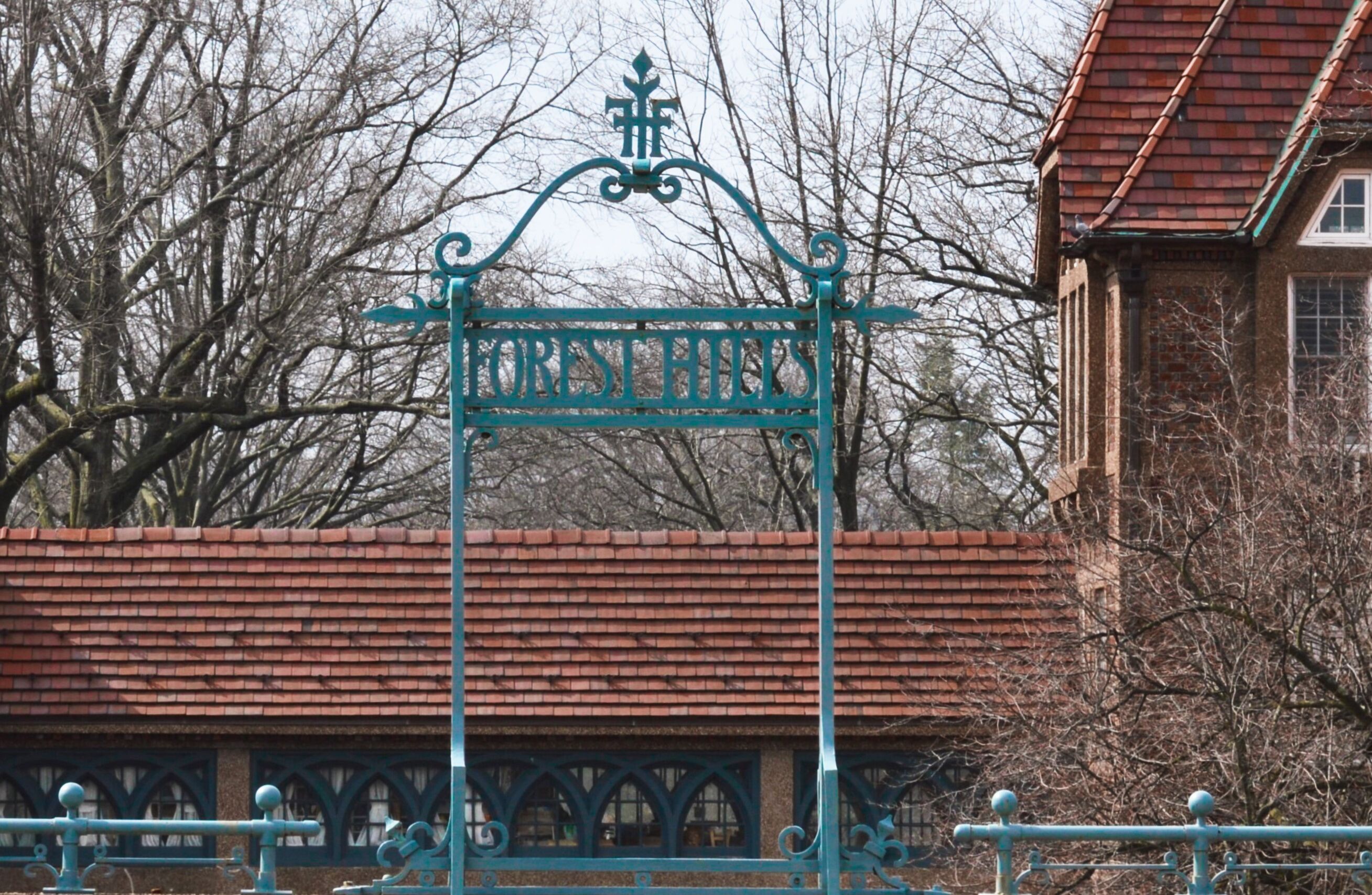 A wrought iron gate with 'FOREST HILLS' inscribed, a brick building, and leafless trees in the background.