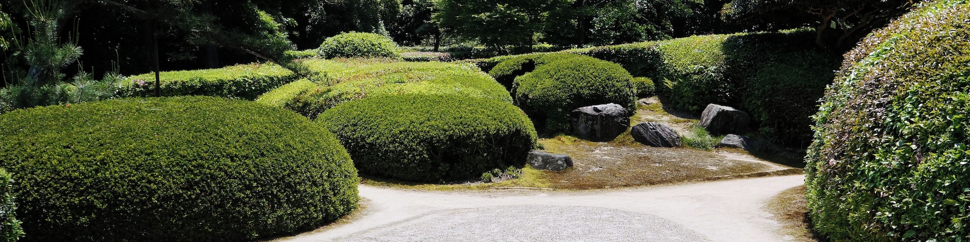 Uma trilha de cascalho serpenteia por um jardim bem cuidado, com arbustos podados e pedras grandes.