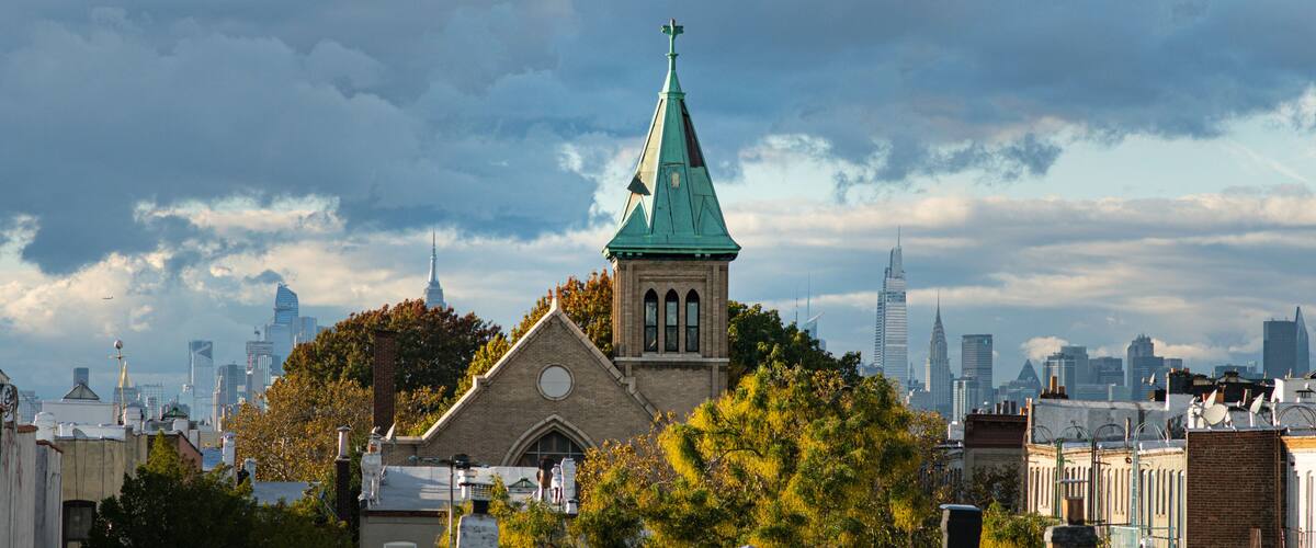 A church with a green spire, surrounded by trees and buildings in a cityscape.