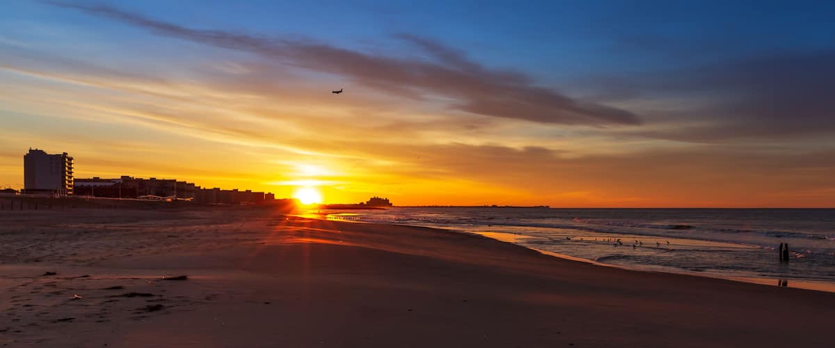 A beach at sunset with the sun low on the horizon, waves gently lapping the shore, and a city skyline in the background.