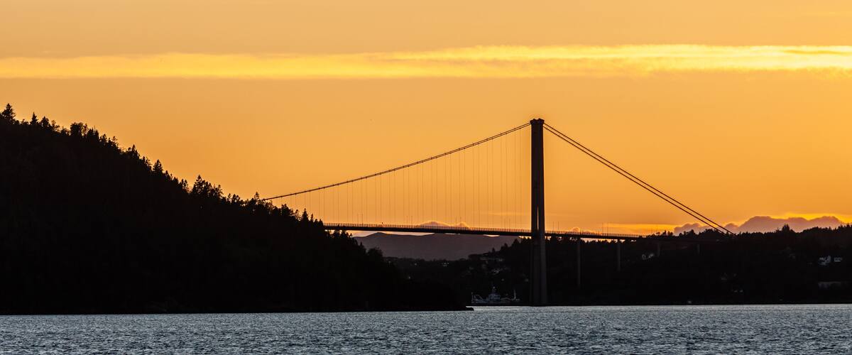 A suspension bridge silhouetted against a sunset sky, with a forested hillside and calm water in the foreground.