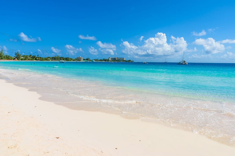Uma praia com águas turquesa cristalinas, areia branca e um veleiro ao longe.