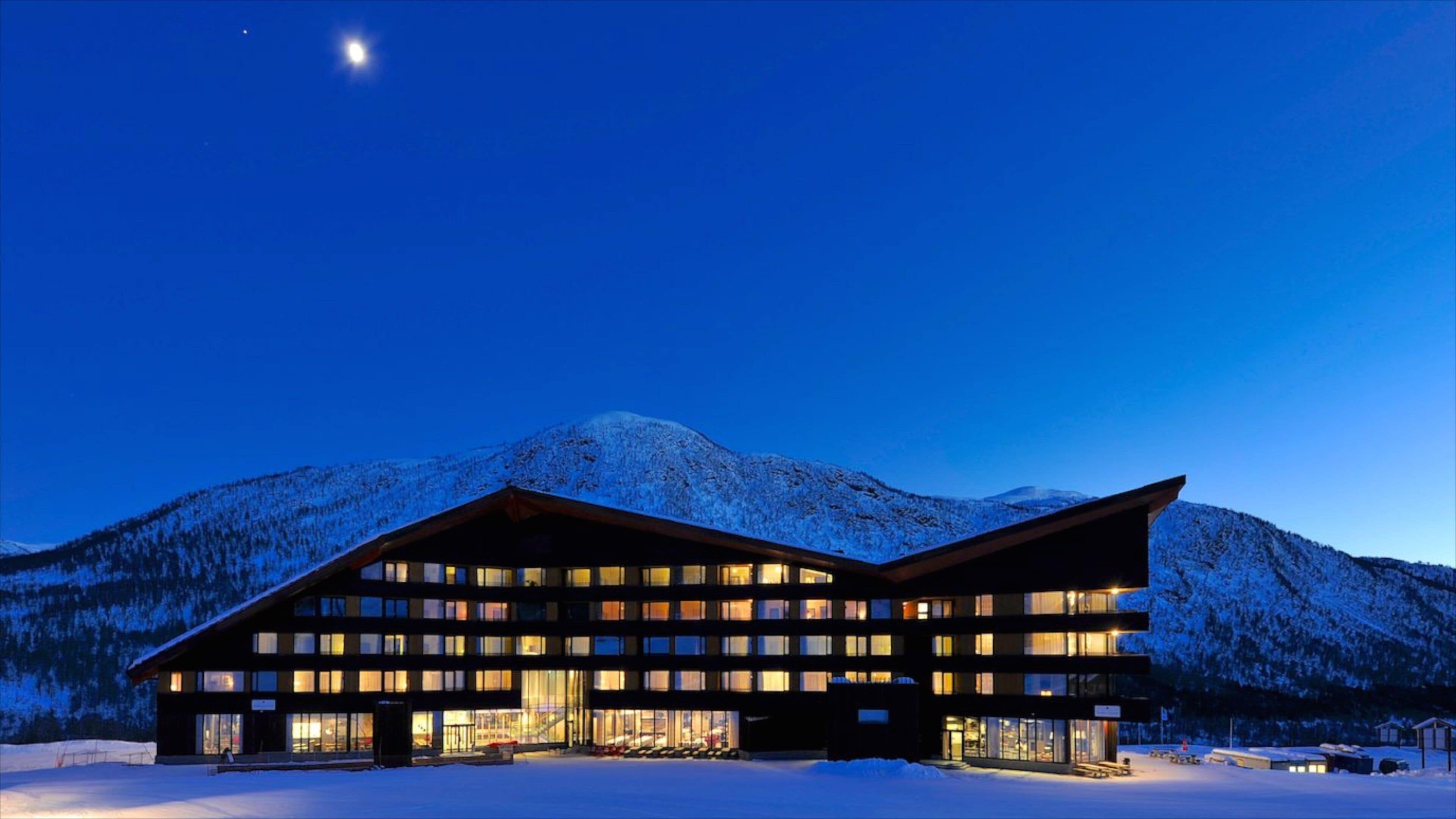 Un edificio moderno con múltiples ventanas y techo inclinado, rodeado de montañas nevadas y un cielo nocturno despejado.