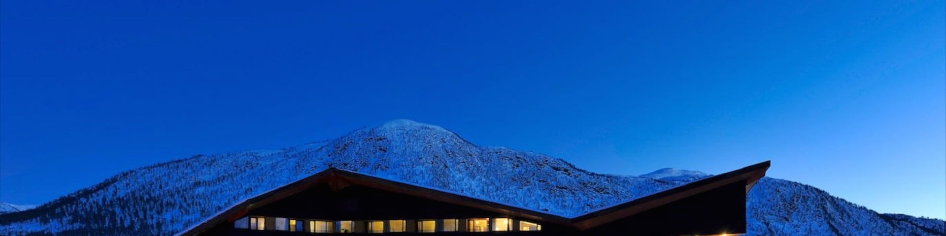 Un edificio moderno con múltiples ventanas y techo inclinado, rodeado de montañas nevadas y un cielo nocturno despejado.
