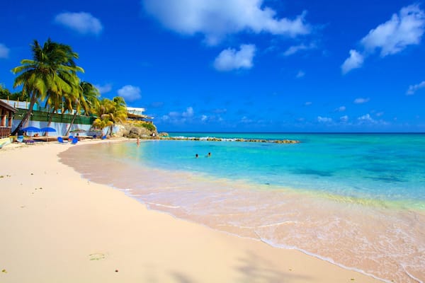 Une plage aux eaux turquoise et limpides, bordée de palmiers, avec un promontoire rocheux qui se dessine à l’horizon.