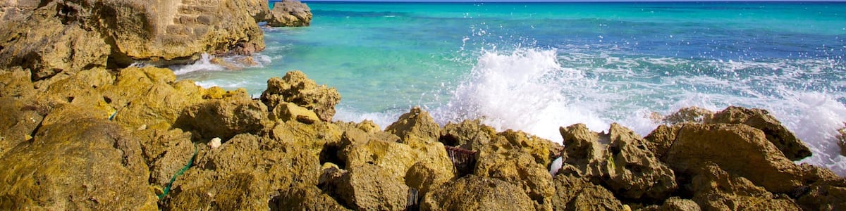 A rocky coastline with clear turquoise waters, a yellow building with a balcony overlooking the sea, and a clear blue sky with scattered clouds.