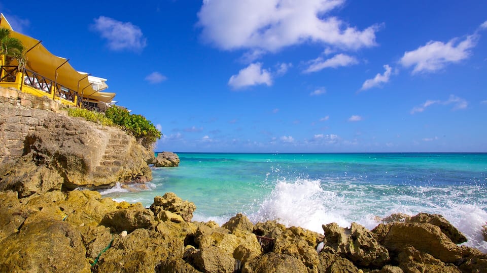 A rocky coastline with clear turquoise waters, a yellow building with a balcony overlooking the sea, and a clear blue sky with scattered clouds.