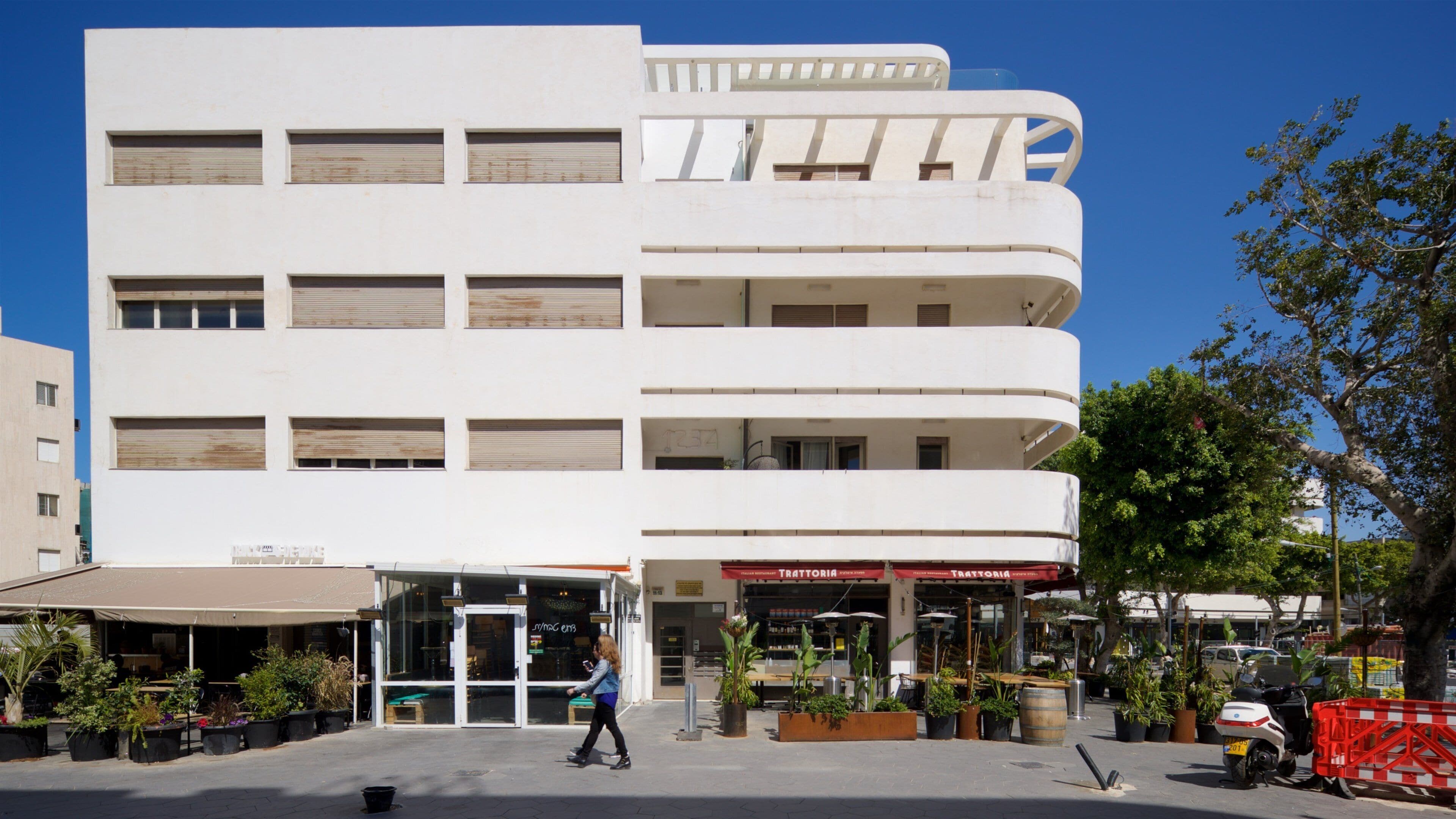 Un immeuble blanc à plusieurs étages, doté de balcons et abritant un commerce de détail appelé « Trattoria ».