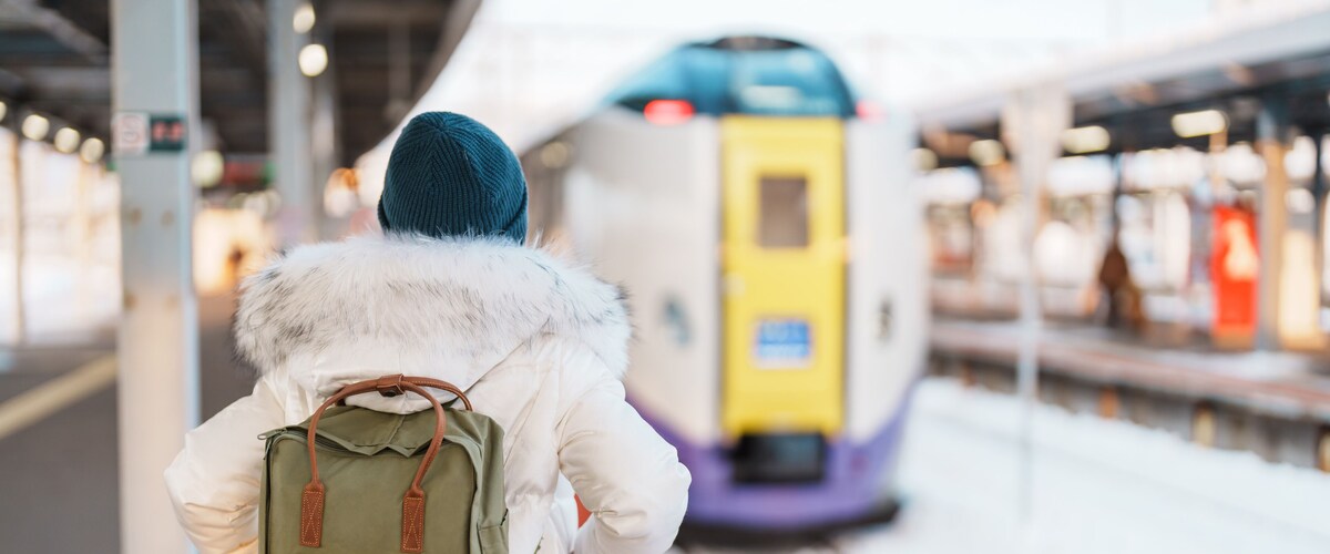 A person with a fur-lined coat and a backpack at a train station with a train in the background.