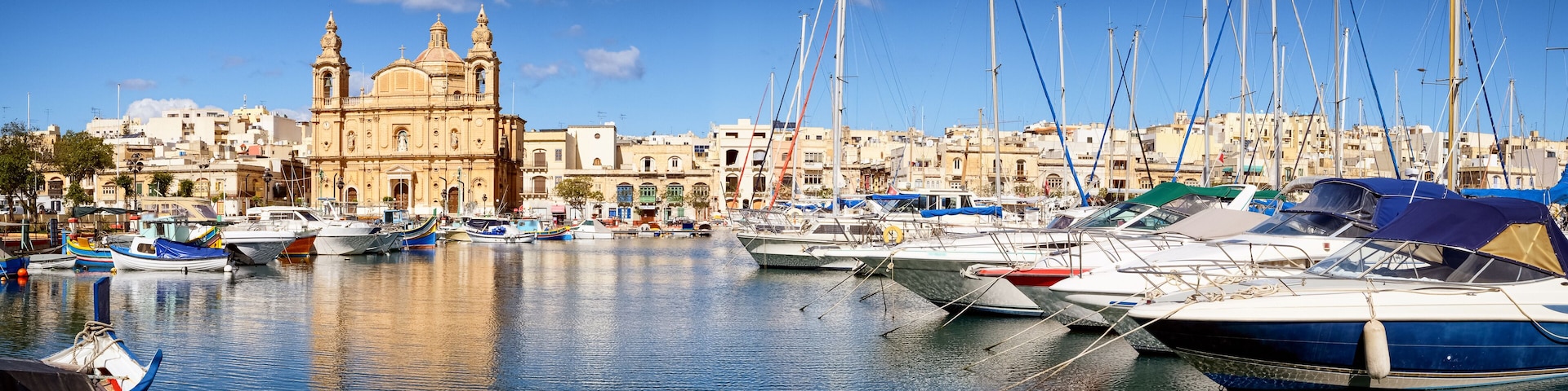 A marina with various boats docked, a prominent church with two towers, and a backdrop of residential buildings.
