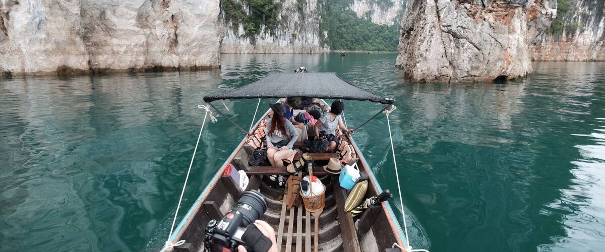 A narrow boat with passengers and a photographer on a calm river surrounded by rocky cliffs.