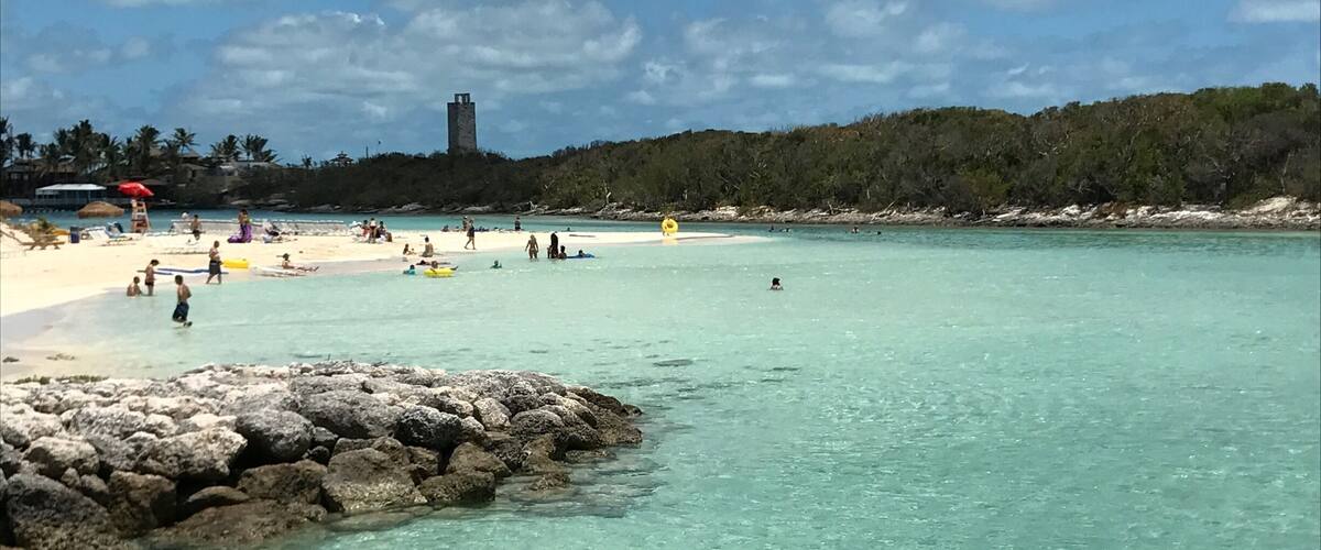 A beach with clear turquoise water, a rocky shoreline, and a distant tower on a hill.