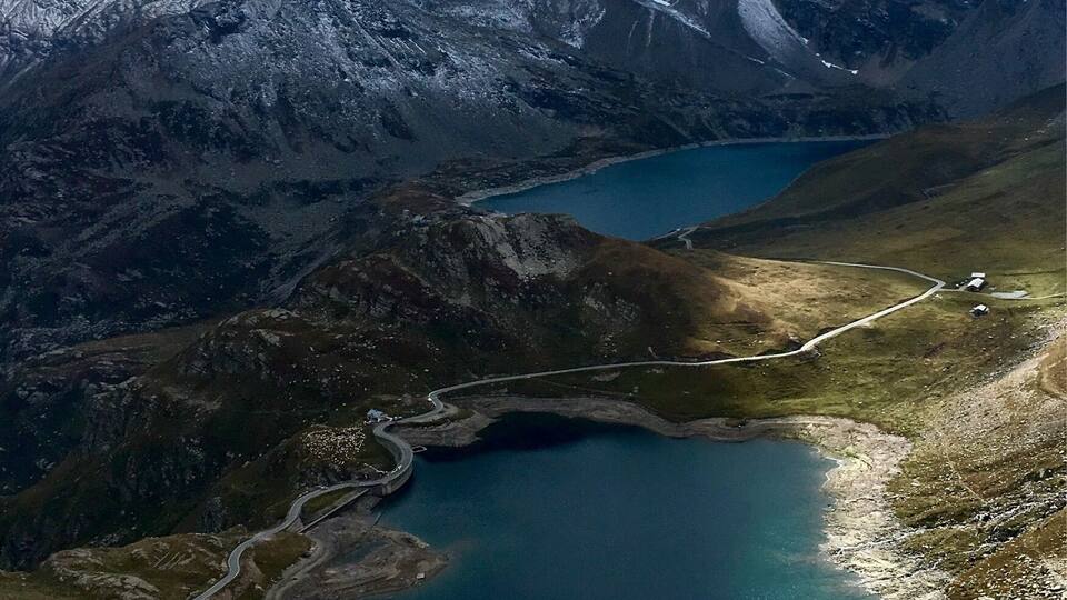 A mountainous landscape with a turquoise lake, a winding road, and snow-capped peaks.
