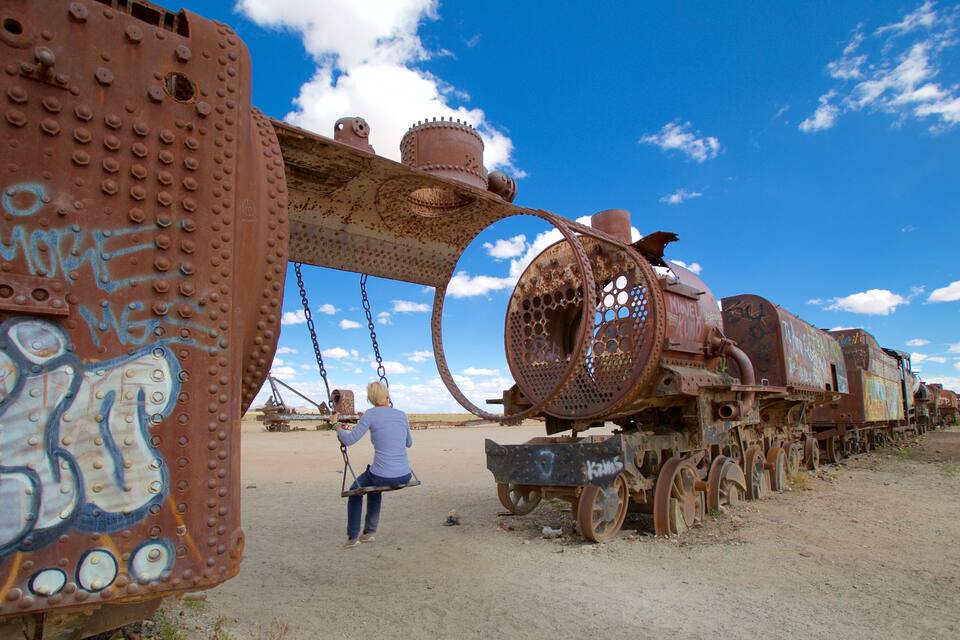 Una locomotiva a vapore arrugginita con graffiti, cielo azzurro limpido e una persona che spinge una carriola.