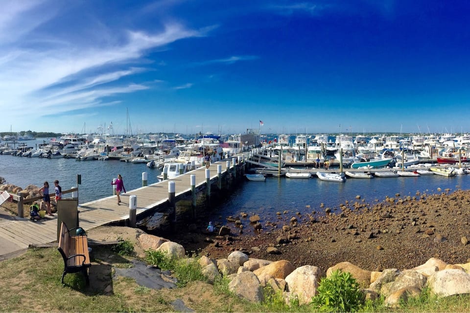 A marina with boats docked along a wooden pier, a rocky shoreline, and a clear blue sky.