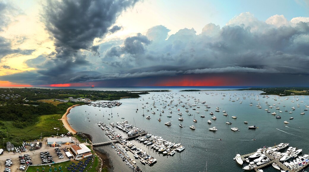 Aerial view of a harbor filled with boats under a dramatic sky at sunset, contrasting with lush green landscapes, New Shoreham, Rhode Island, United States.