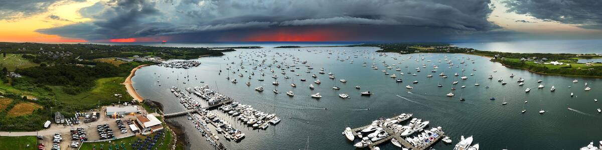 Aerial view of a harbor filled with boats under a dramatic sky at sunset, contrasting with lush green landscapes, New Shoreham, Rhode Island, United States.