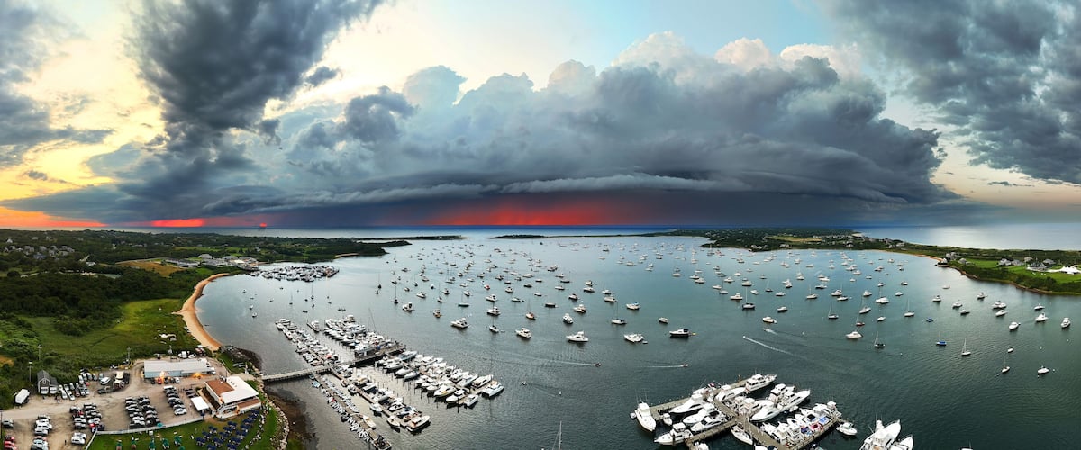 Aerial view of a harbor filled with boats under a dramatic sky at sunset, contrasting with lush green landscapes, New Shoreham, Rhode Island, United States.