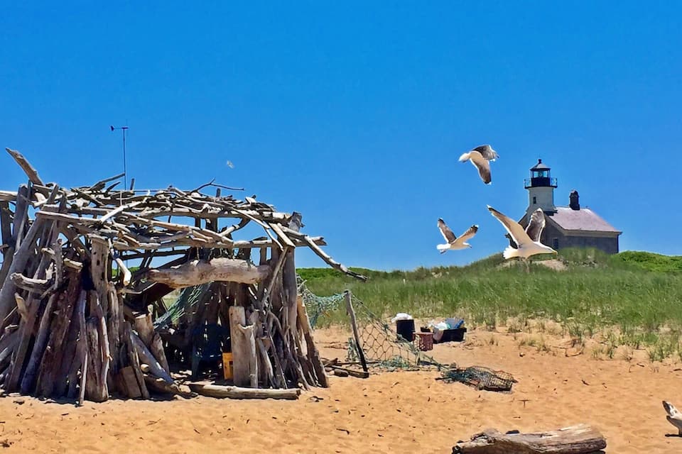 Una playa arenosa con una estructura de madera, un faro y gaviotas volando.