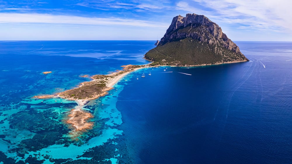 Luftaufnahme einer Küsteninsel mit klarem, blauem Wasser, einem zerklüfteten Berg und einer schmalen Halbinsel mit deutlichen Bootsspuren.