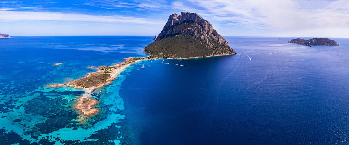 An aerial view of a coastal island with clear blue waters, a rugged mountain, and a narrow peninsula with visible boat tracks.
