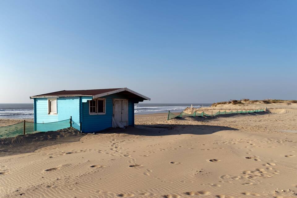 A small blue beach hut on a sandy beach with footprints leading to it.