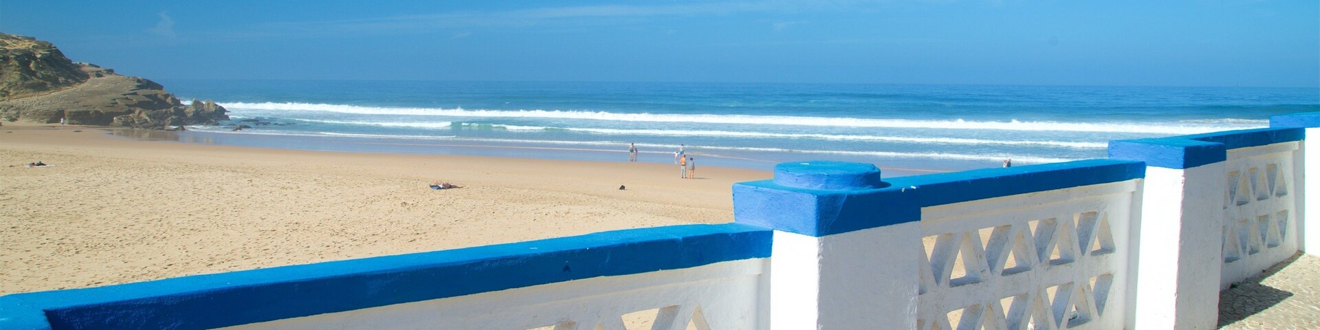 Una playa con una cerca blanca y azul, orilla arenosa y olas oceánicas.