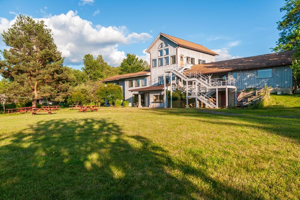 A large wooden house with a staircase leading to a deck, surrounded by a grassy lawn and trees.