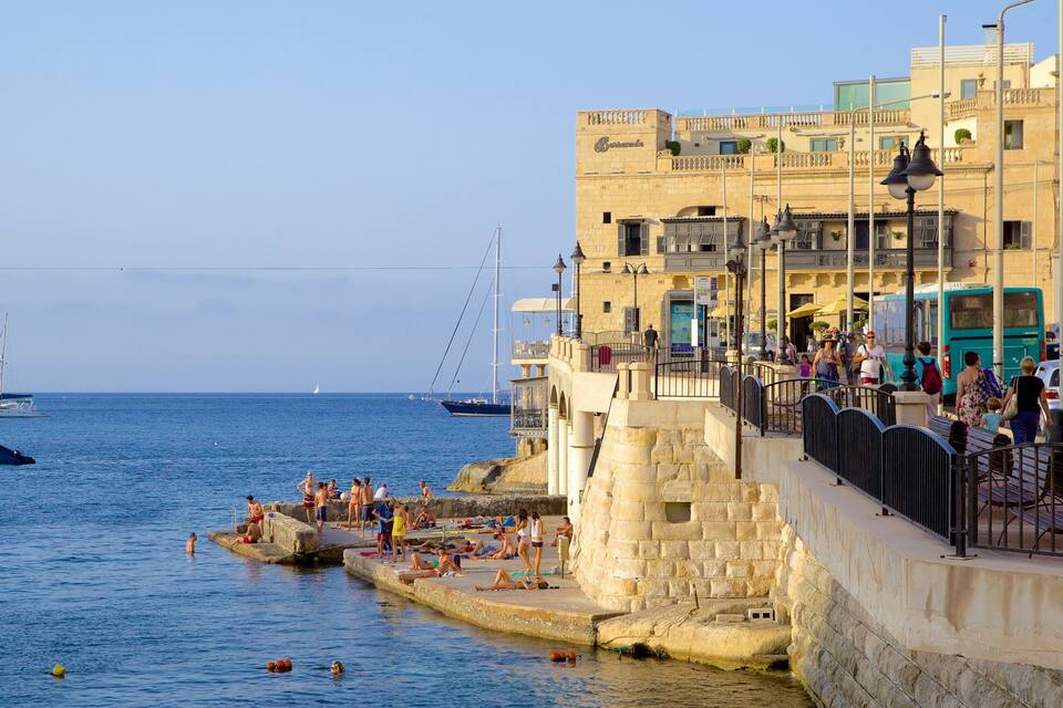 Un paseo marítimo costero con gente relajándose junto al mar, un muro de piedra y un edificio con un letrero.