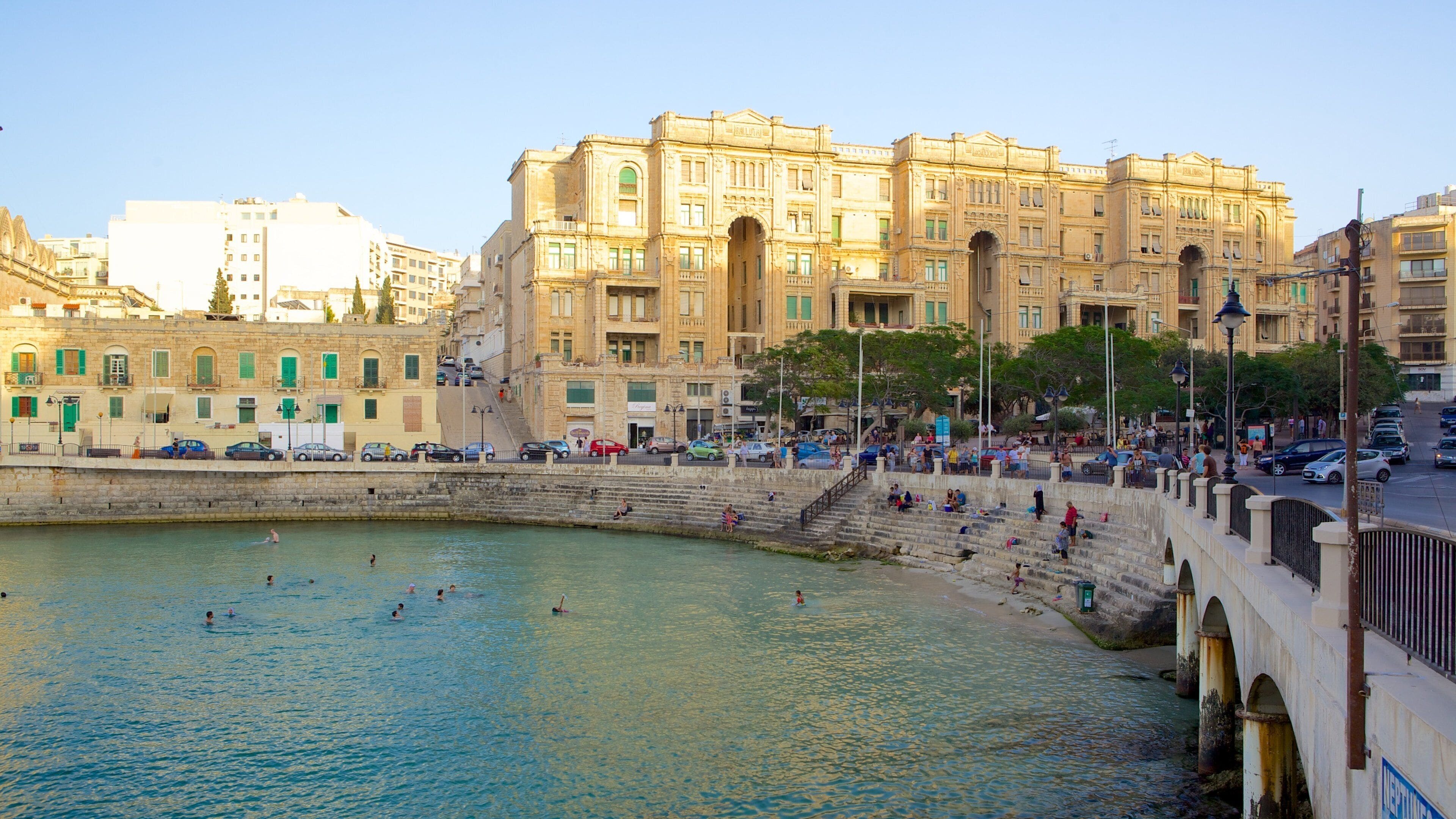 A waterfront promenade with people swimming and relaxing, surrounded by historic buildings.