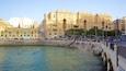 A waterfront promenade with people swimming and relaxing, surrounded by historic buildings.