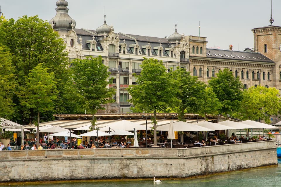 A riverside café with outdoor seating, surrounded by trees and historic buildings.