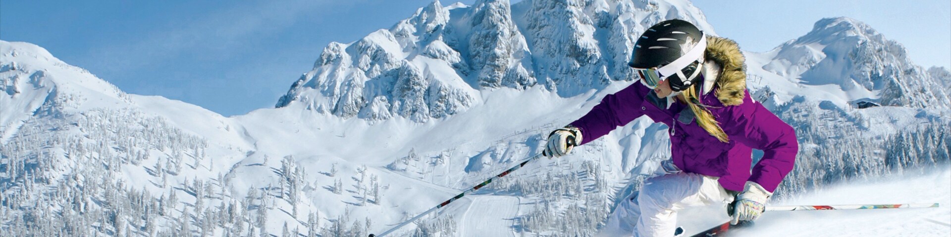 Un esquiador con chaqueta morada y pantalones blancos esquía por una pendiente nevada, con árboles cubiertos de nieve y montañas al fondo.