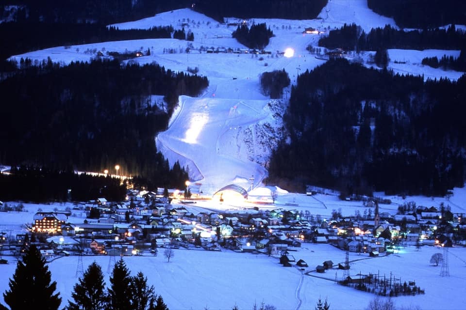 Une station de ski enneigée, illuminée la nuit, avec des bâtiments éclairés et un télésiège.