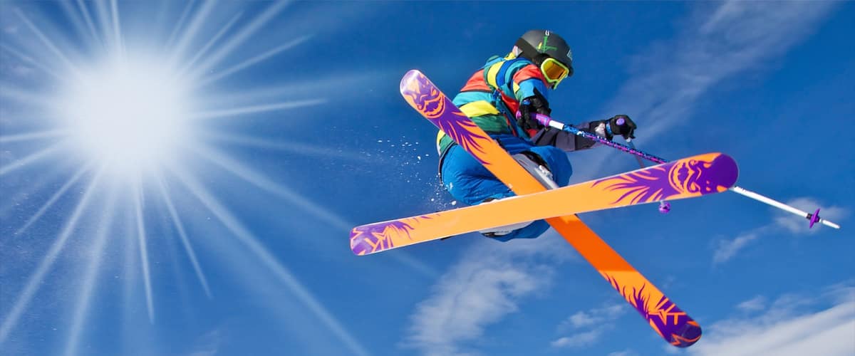 A skier in mid-air with skis crossed, against a clear blue sky and bright sun.