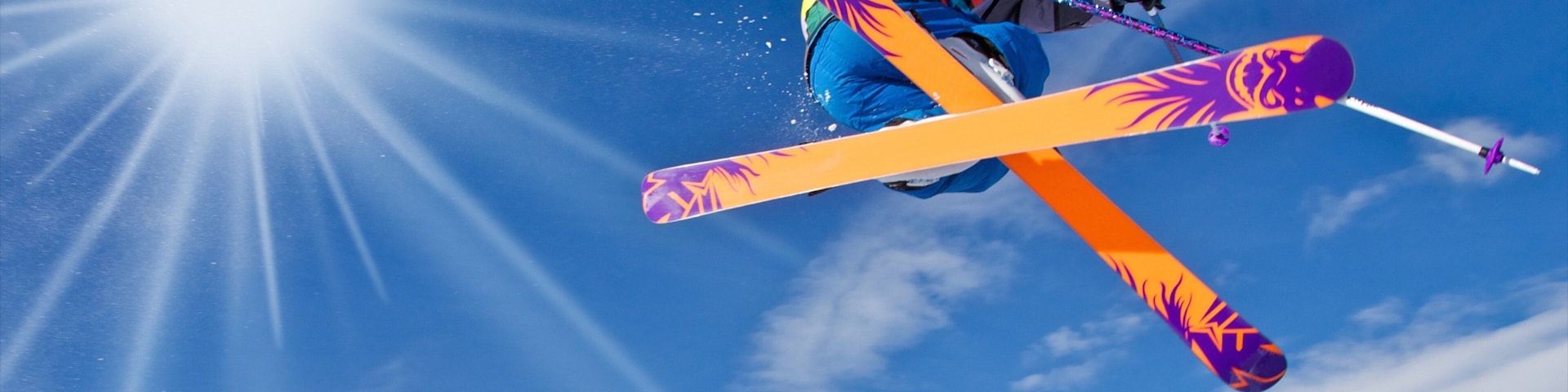 A skier in mid-air with skis crossed, against a clear blue sky and bright sun.