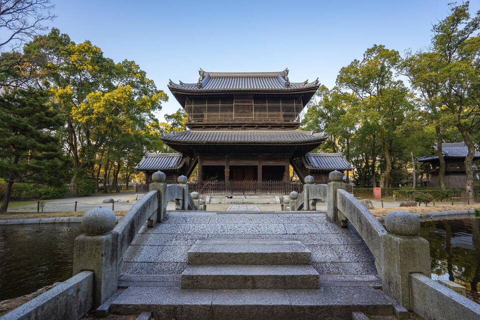Um templo japonês tradicional com uma ponte de pedra que leva até ele, cercado por árvores e um lago.